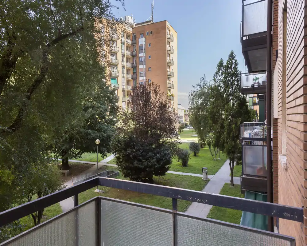 Balcone soleggiato con vista su un cortile verde con alberi e palazzine vicine — ideale per un caffè mattutino o per rilassarsi all’aperto.