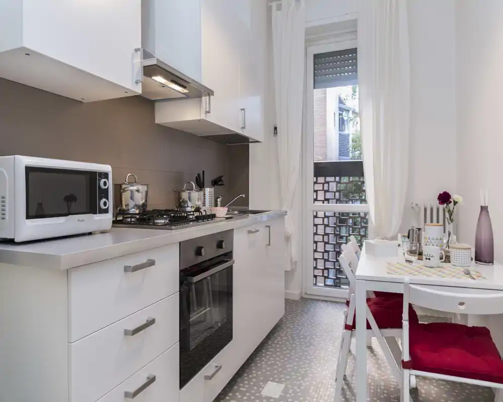 Cucina compatta e moderna con mobili bianchi, forno incassato e piano cottura a gas, piccolo tavolo con cuscini rossi e porta sul balcone — pratica e ben arredata per la vita in città.
