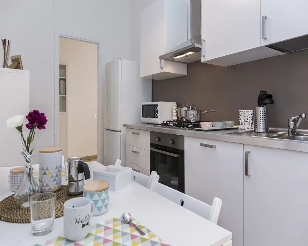 Cucina moderna e compatta con mobili bianchi, forno integrato, lavello e tavolo apparecchiato con tovagliette colorate e barattoli decorativi — luminosa e accogliente, ideale per affitti brevi.