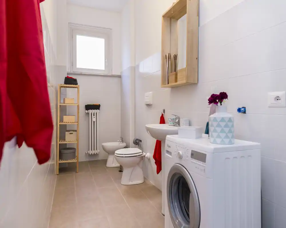 Bagno moderno e luminoso con lavatrice, lavabo, WC e bidet, scaffalatura in legno e dettagli rossi.