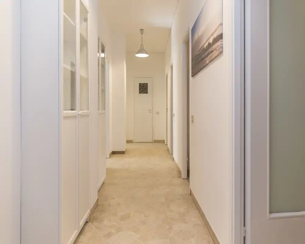 Corridoio luminoso e pulito con pareti neutre, armadi a sinistra e un lampadario a sospensione che conduce alle porte interne — perfetto come ingresso.