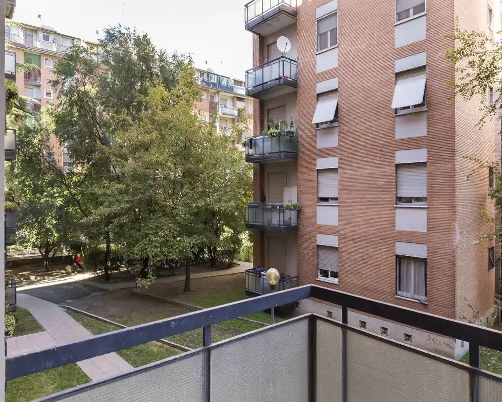 Vista dal balcone soleggiato su un cortile alberato con percorsi e palazzine di mattoni vicine.