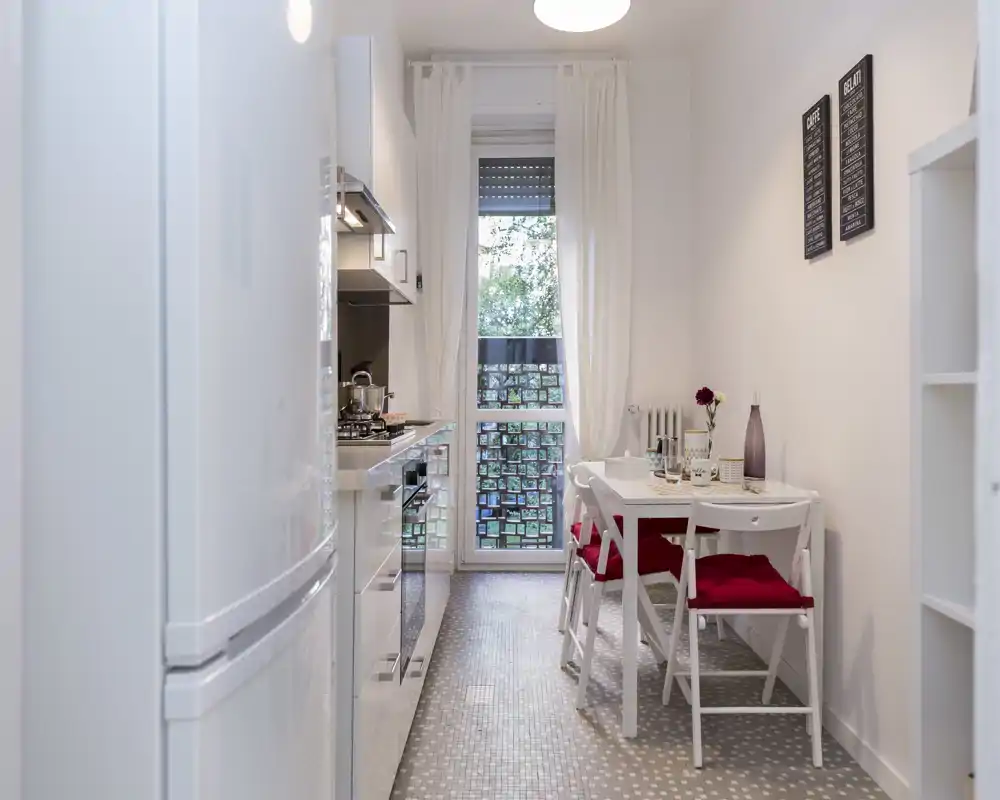 Cucina stretta e luminosa in stile corridoio con mobili bianchi, piccolo tavolo da pranzo e cuscini rossi. Lo spazio è pulito, ben illuminato e rifinito con dettagli decorativi semplici.