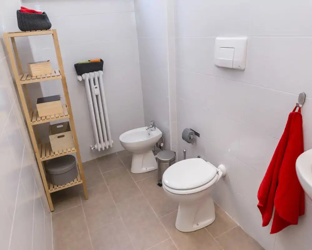 Bagno compatto e pulito con wc, bidet, lavabo parzialmente visibile e una scaffalatura in legno per riporre oggetti. Piastrelle chiare e un asciugamano rosso donano un tocco di colore.