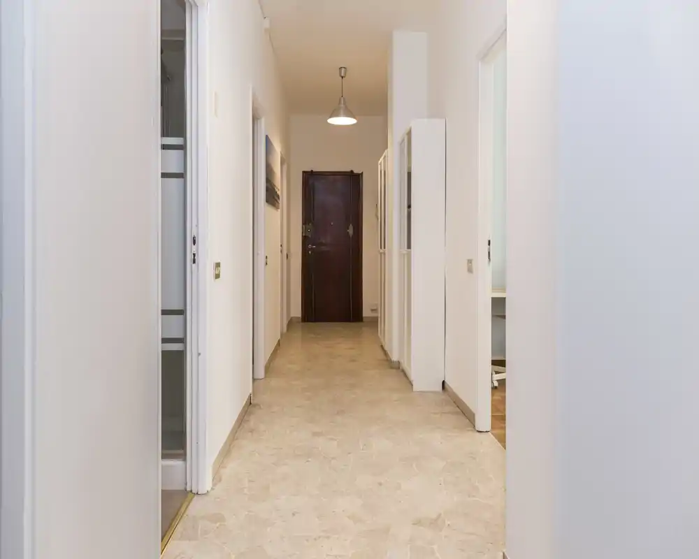 Ingresso/corridoio luminoso e ordinato con pavimento in piastrelle, pareti bianche e porta d’ingresso in legno sul fondo. Il corridoio è ben illuminato e mostra le porte delle stanze adiacenti.
