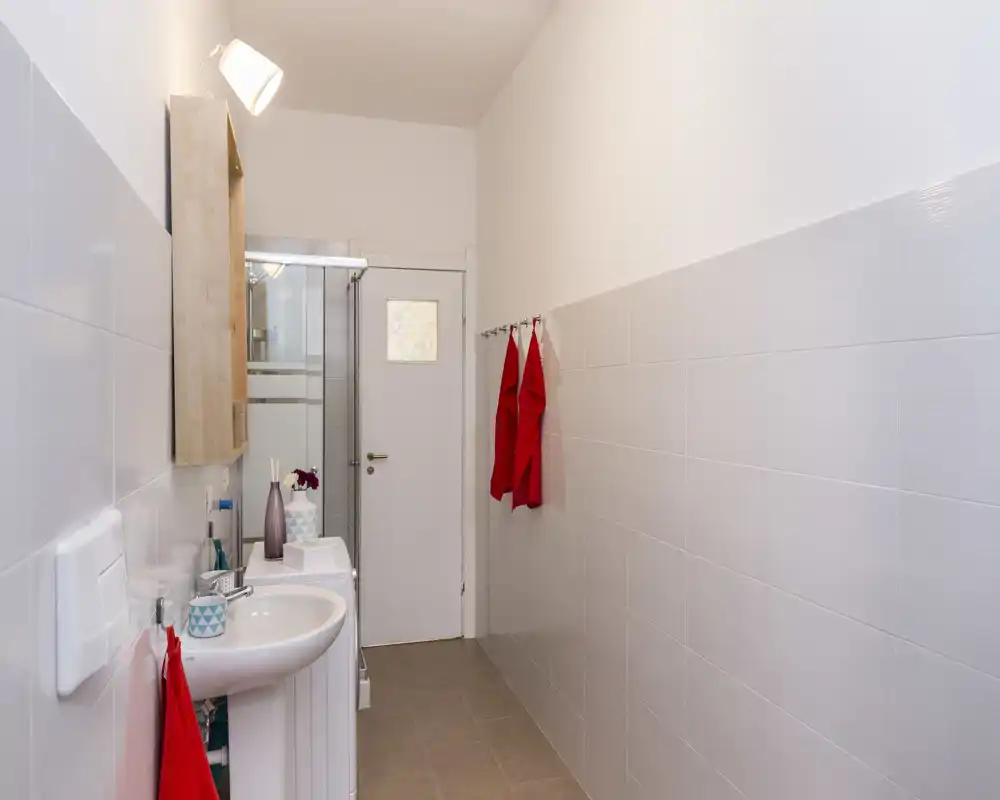 Bagno moderno e stretto con lavabo, specchio con pensile, box doccia e asciugamani appesi; piastrelle bianche luminose e stile minimal pulito.