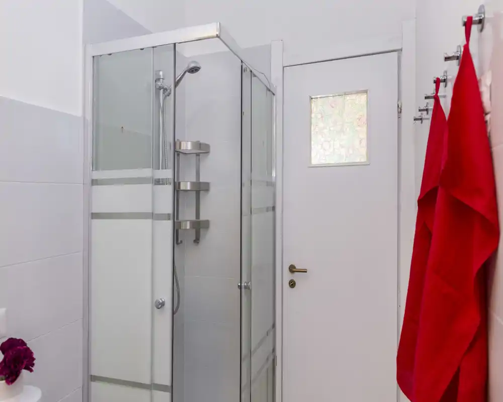 Bagno moderno e pulito con box doccia angolare in vetro, piastrelle bianche e asciugamani rossi appesi.