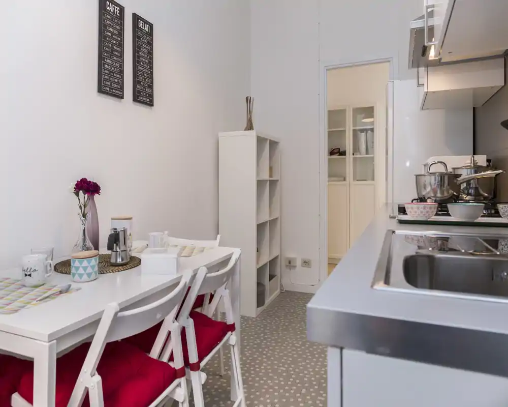 Cucina moderna compatta con lavello in acciaio e piano cottura sul lato destro e un piccolo tavolo da pranzo a sinistra con sedie con cuscini rossi, decorata con tovagliette e un vaso di fiori.