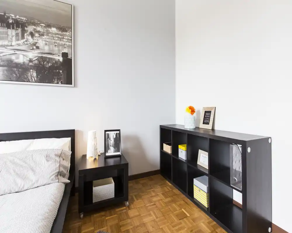 Dormitorio luminoso y minimalista con cama doble, mesita de noche y estantería abierta; líneas limpias y luz natural que aportan sensación de amplitud.