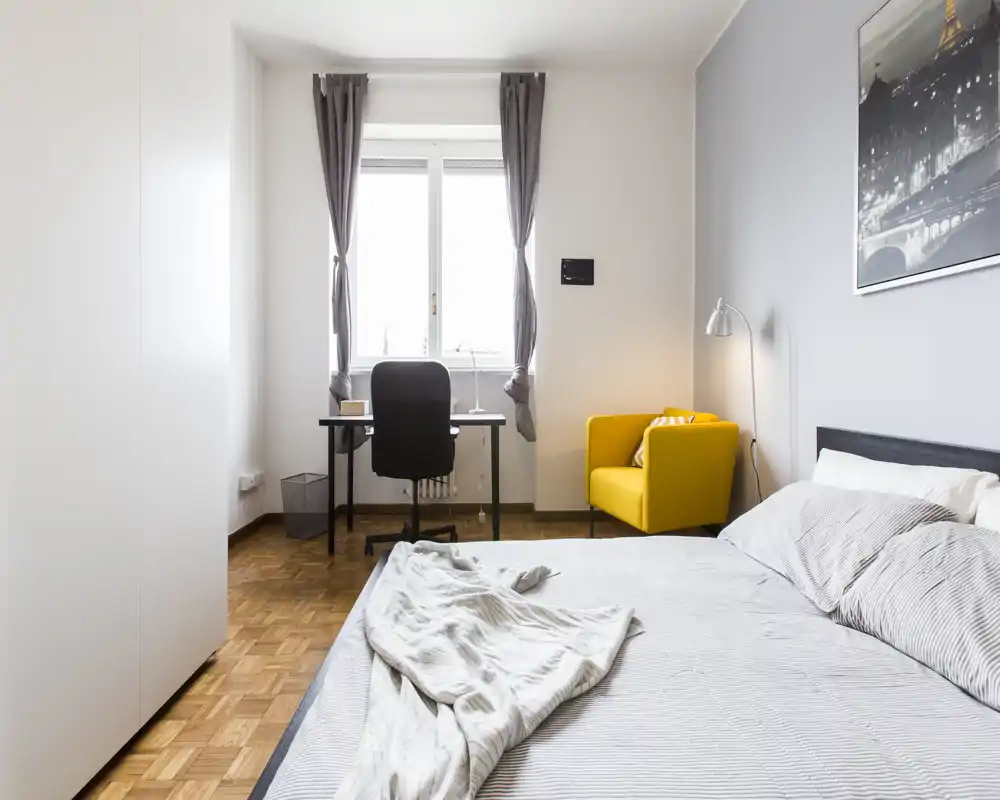 Dormitorio moderno y luminoso con una cama grande en primer plano, un escritorio frente a la ventana y un sillón amarillo. Líneas limpias, luz natural y textiles acogedores hacen la estancia atractiva.