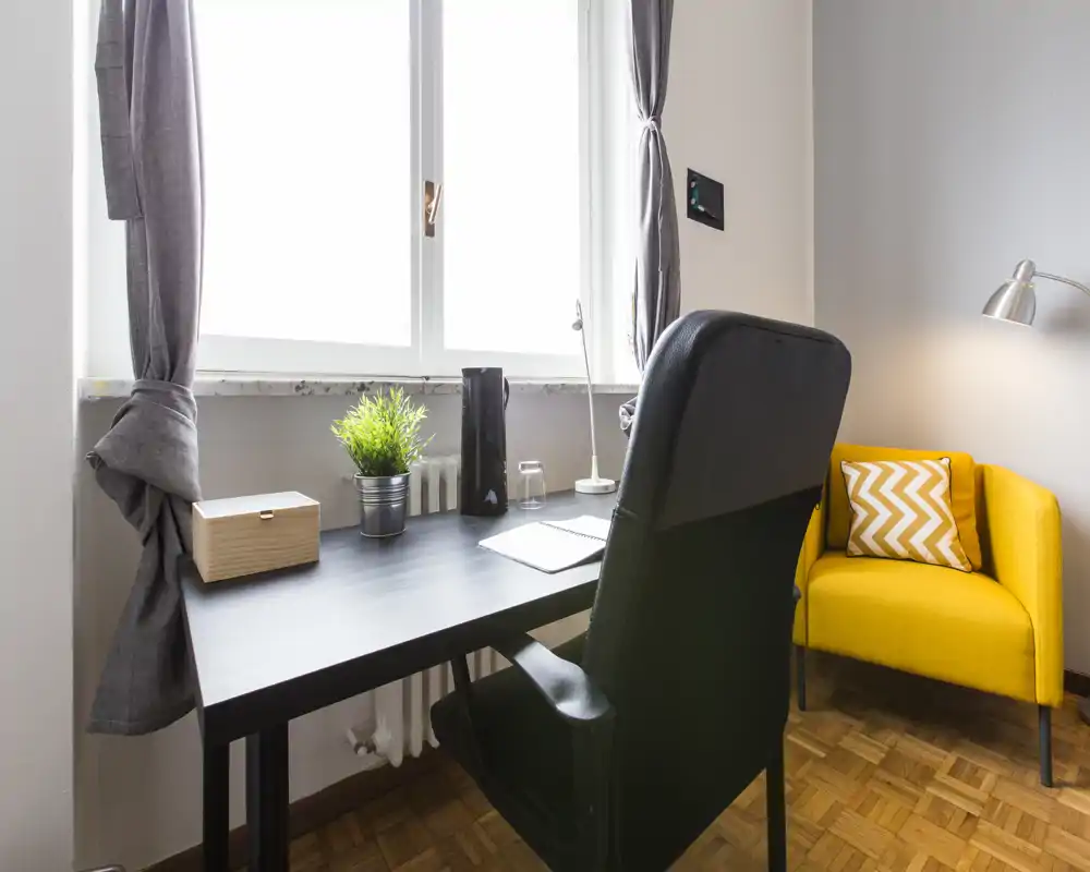Zona de estar luminosa y ordenada con un escritorio junto a la ventana y un acogedor sillón amarillo — ideal como rincón de trabajo en casa.