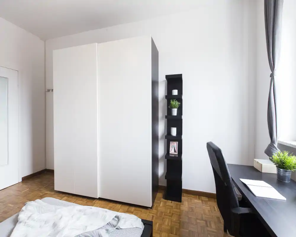 Dormitorio luminoso y minimalista con un gran armario blanco, estantería lateral y un escritorio junto a la ventana; cama parcialmente visible en primer plano con ropa de cama suave.