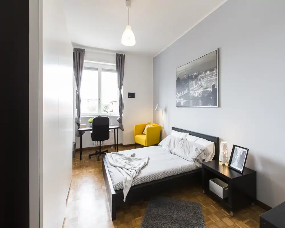 Dormitorio moderno y luminoso con cama doble, mesita, escritorio junto a la ventana y un sillón amarillo como punto focal; ambiente limpio y acogedor.