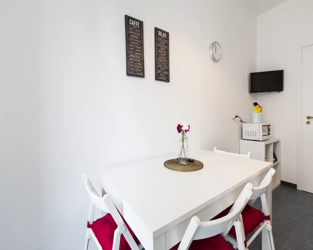 Acogedor rincón de comedor luminoso y minimalista con mesa blanca, cuatro sillas con cojines rojos, un pequeño centro de mesa y decoración en la pared — ideal para pisos compactos.
