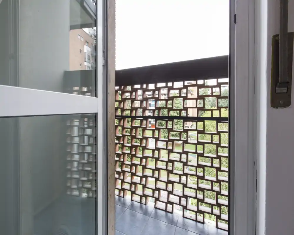 Pequeño balcón visto a través de una puerta corredera, con un panel decorativo de ladrillo y suelo de baldosas, que ofrece vistas parciales a un patio ajardinado.