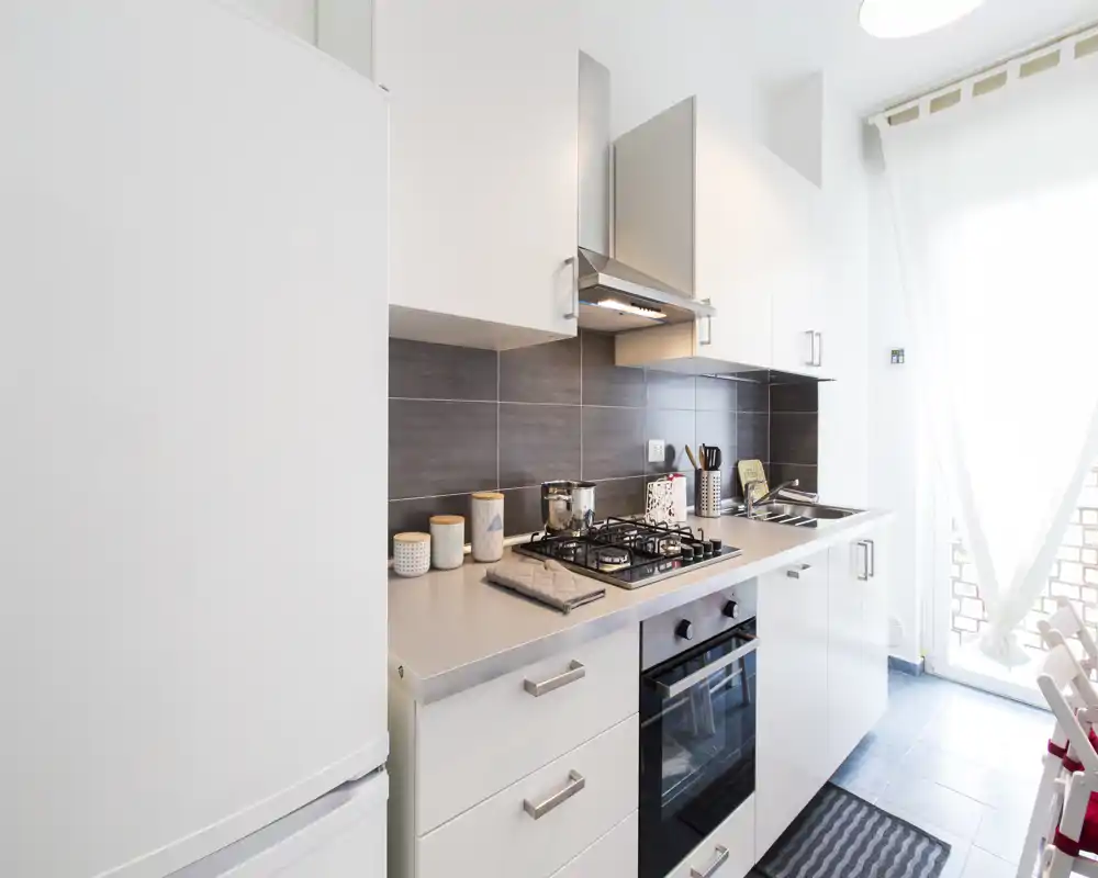Cocina moderna y compacta con placa de gas, horno empotrado, armarios blancos y una ventana/puerta luminosa que aporta luz natural. Encimera despejada con pequeños frascos decorativos y utensilios organizados.