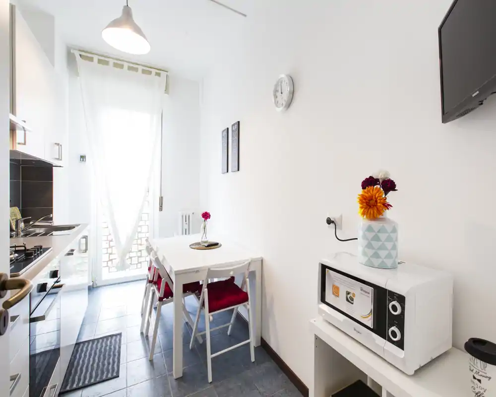 Cocina compacta y luminosa con mesa pequeña, cocina de gas, microondas y luz natural desde una puerta acristalada — ideal para un apartamento urbano acogedor.