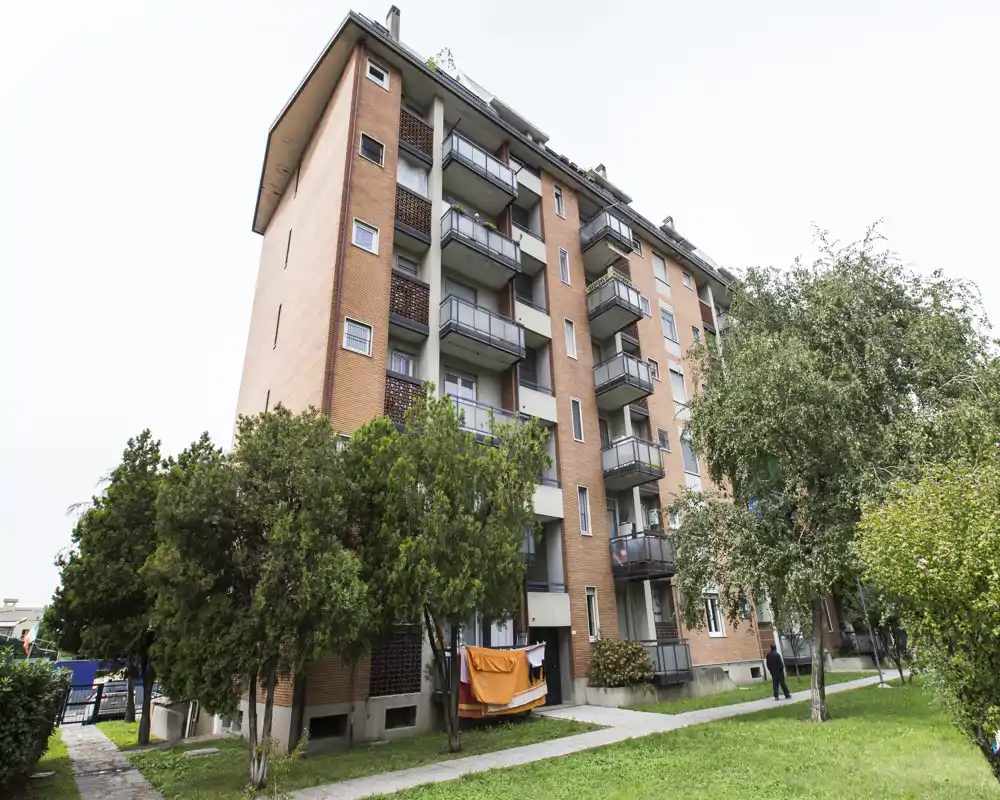 Vista exterior de un edificio residencial de varias plantas con balcones, césped y árboles; se aprecia el área exterior comunal.