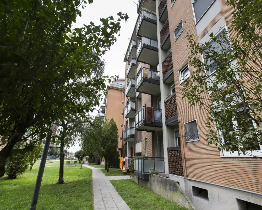 Vista exterior de un edificio residencial con balcones, una senda pavimentada y césped con árboles — ideal para mostrar la fachada y el acceso exterior.