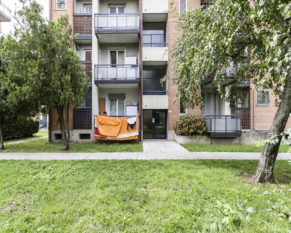 Fachada exterior de un edificio residencial con pequeños balcones, un balcón en planta baja con ropa tendida y un área común con césped al frente.