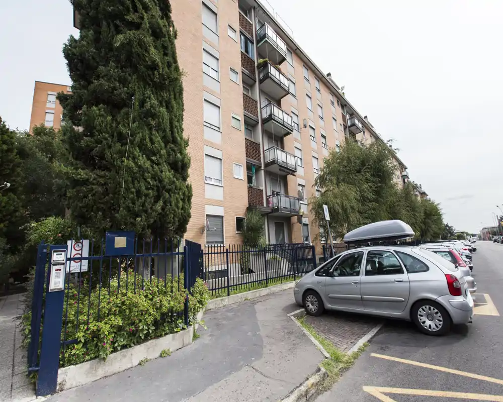 Vista exterior de un edificio residencial con entrada vallada, aparcamiento en la calle y balcones — útil para mostrar la fachada y el entorno del vecindario.