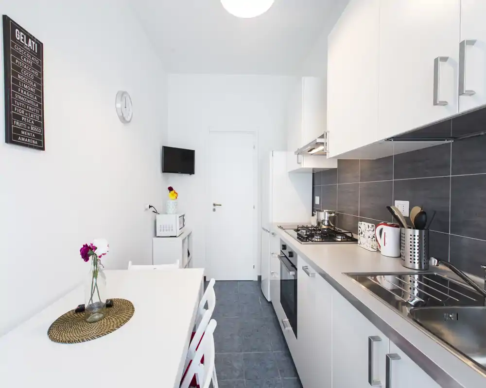 Cocina moderna y luminosa con armarios blancos, fregadero de acero y vitro a gas; una mesa pequeña con sillas y flores decorativas crean un espacio práctico y acogedor para cocinar y comer.