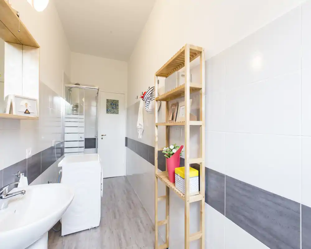 Baño moderno y luminoso con lavabo, lavadora y ducha. Azulejos neutros y estantería de madera ofrecen una imagen limpia y funcional, ideal para alquileres.