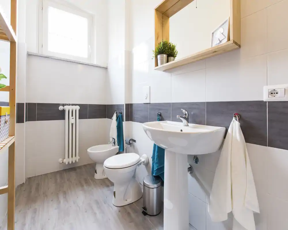 Baño moderno y luminoso con lavabo de pedestal, inodoro, bidé y suelo efecto madera; azulejos blancos con franja horizontal oscura y una pequeña ventana que aporta luz natural.