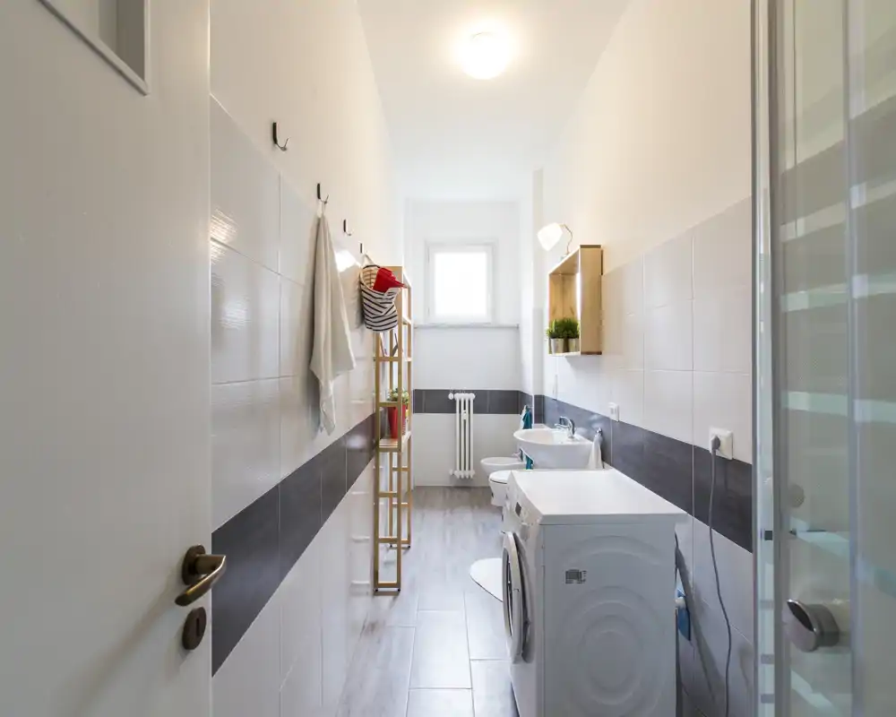 Baño largo y luminoso con ducha, lavabo, inodoro y lavadora — azulejos modernos y luz natural desde la ventana.