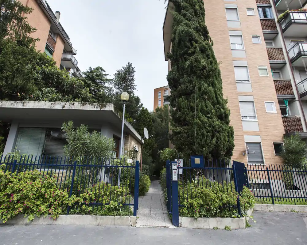 Entrada principal y pequeño jardín de un edificio con acceso con valla, paseo y árboles maduros—luminoso, verde y bien cuidado.