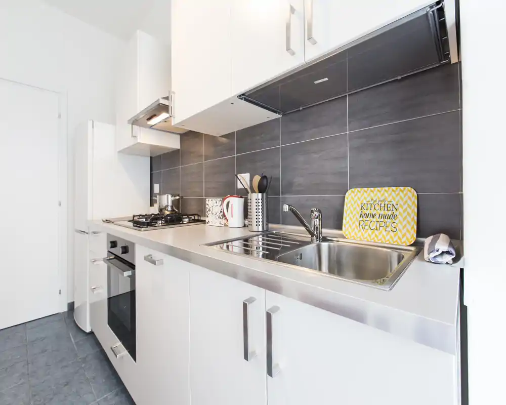 Cocina moderna y compacta con fregadero de acero inoxidable, placa de gas y muebles blancos. Superficies limpias y luminosas y accesorios cuidados que crean un espacio de cocina práctico y con estilo.