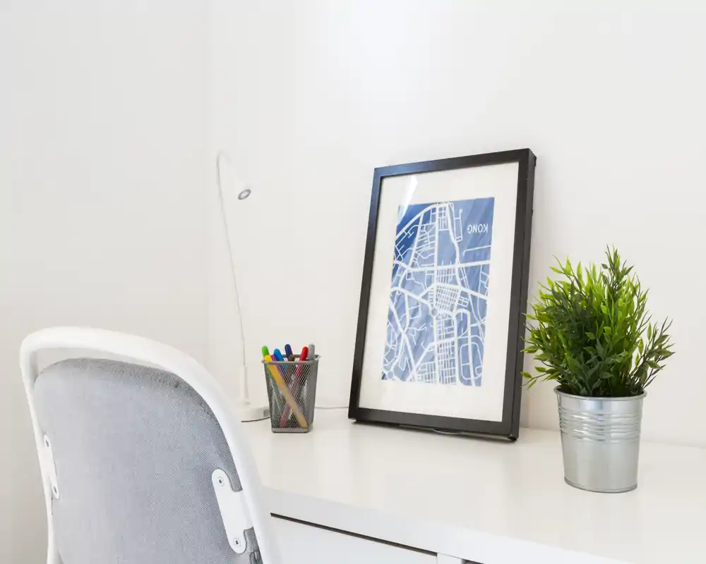 Piccolo angolo scrivania minimal e luminoso con una stampa incorniciata, una pianta in vaso e un portapenne su una superficie bianca — perfetto per foto di home office.