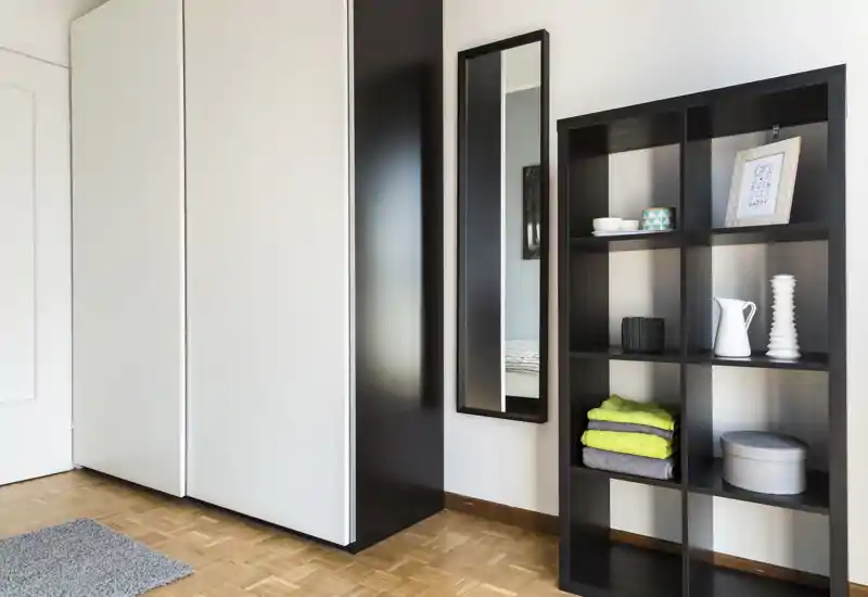 Rincón de dormitorio ordenado con un gran armario, espejo de pared y estantería abierta con toallas dobladas y objetos decorativos; estilo moderno y minimalista con suelo de parquet y buena luz natural.