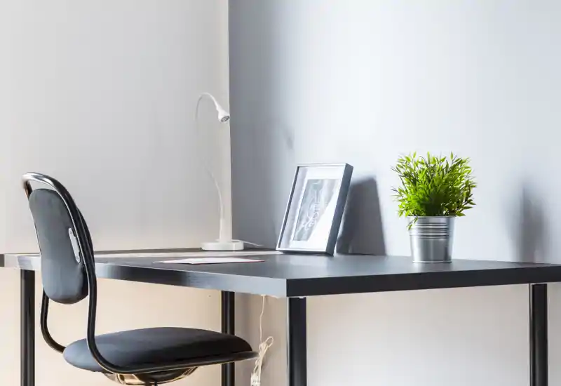 Espacio de trabajo minimalista con escritorio negro, lámpara de mesa, marco decorativo y una pequeña planta en maceta, acompañado de una silla de oficina simple — luminoso y ordenado.