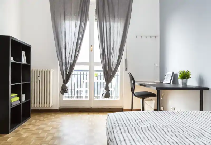 Dormitorio luminoso y moderno con cama bien hecha en primer plano, escritorio junto a la ventana y estantería para almacenamiento: ideal para descansar y trabajar.