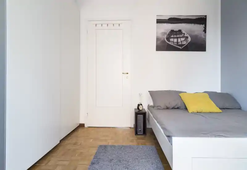 Dormitorio luminoso y minimalista con cama doble en ropa de cama gris, cojín amarillo de acento, mesita y armario — limpio, moderno y acogedor.