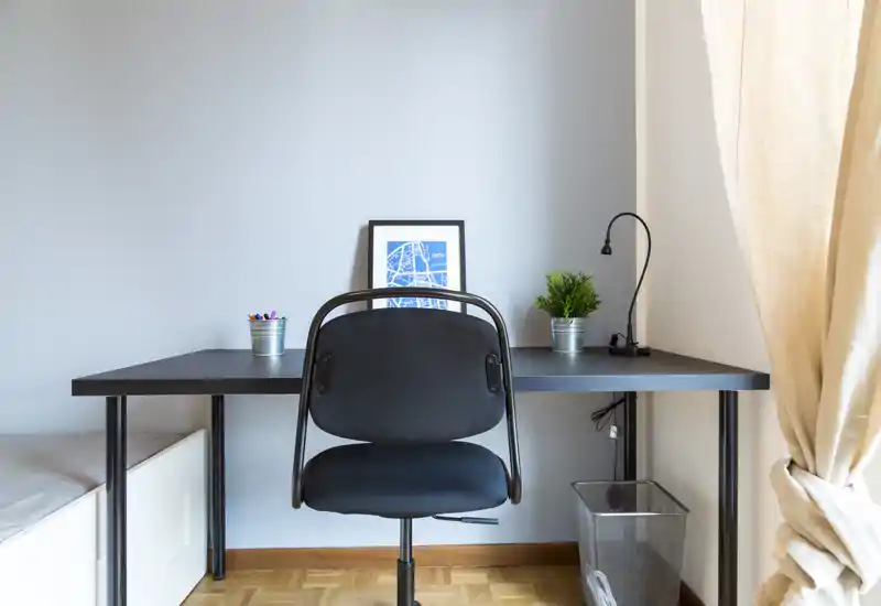 Rincón de trabajo ordenado con escritorio negro y silla ergonómica, acompañado de una pequeña planta y una lámina enmarcada; la luz natural de la ventana aporta un ambiente tranquilo y funcional.
