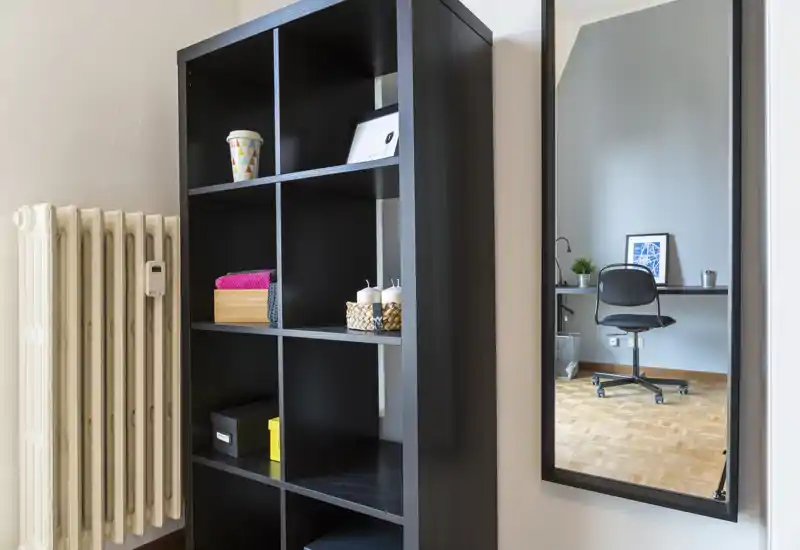 Área de estar de uso múltiple ordenada con una estantería negra y un espejo en la pared que refleja un escritorio y una silla de oficina; tonos neutros y suelo de parquet transmiten una atmósfera funcional y tranquila.