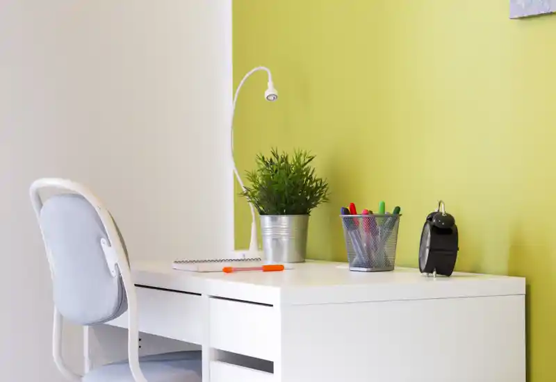 Rincón de estudio luminoso y ordenado con escritorio blanco, silla confortable, lámpara de trabajo, planta en maceta y material de escritura organizado — ideal para teletrabajo o estudio.
