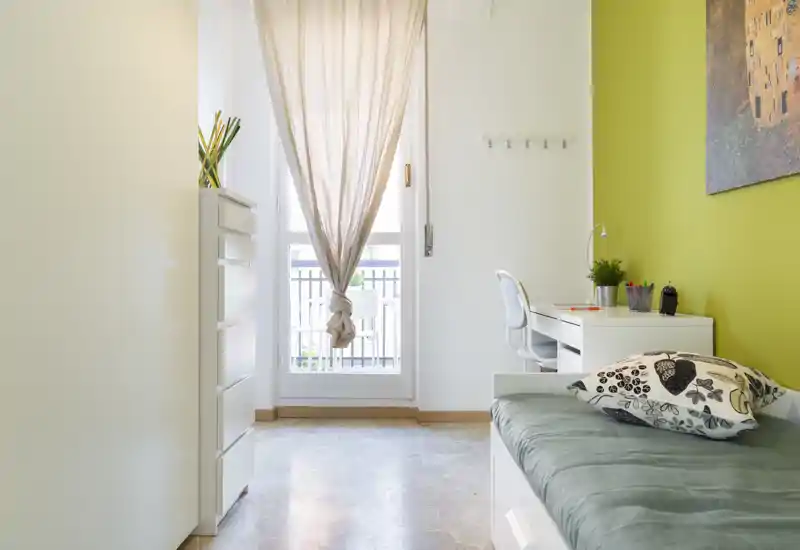 Dormitorio individual luminoso y ordenado con cama tipo día, escritorio y luz natural desde una puerta a un pequeño balcón. Tonos neutros y verdes que crean un ambiente fresco y acogedor, perfecto para alquileres temporales o estudiantes.