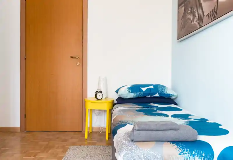 Tidy single bedroom corner with patterned bedding, a bright yellow bedside table and a bedside lamp; simple, well-lit and ready for guests.