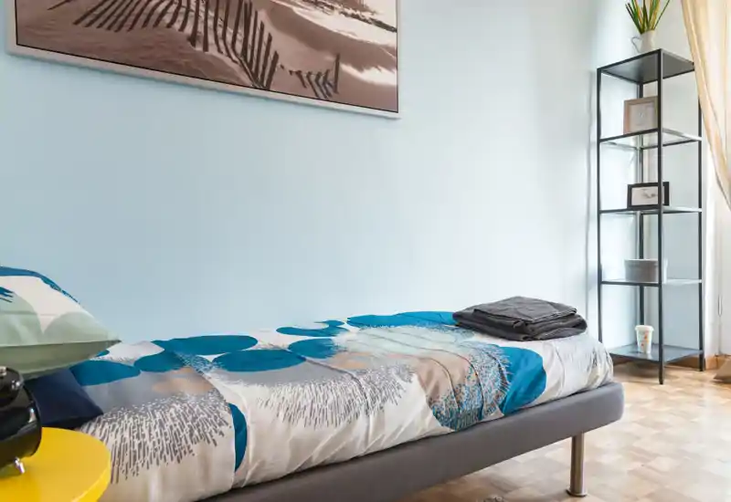 Bright, modern single bedroom with a neatly made bed, colorful duvet and minimalist shelving beside a sunny window.