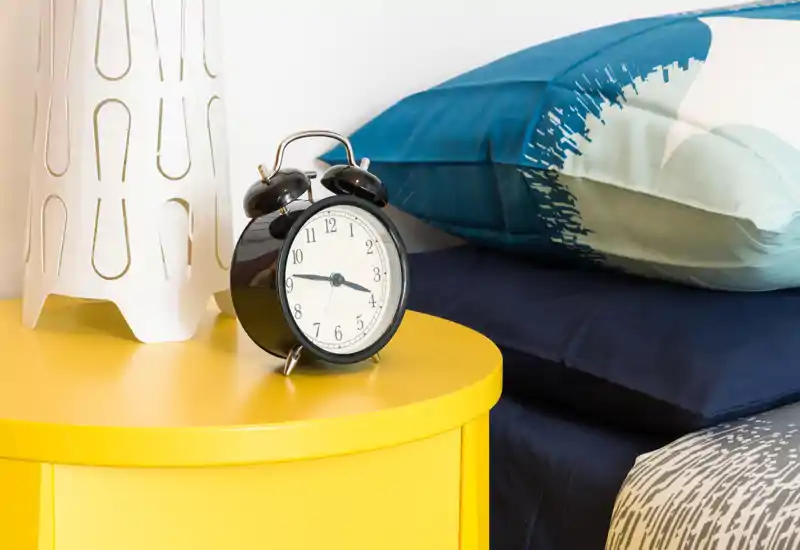 Close-up of a bedroom bedside featuring a retro alarm clock on a bright yellow nightstand, decorative pillows and soft linens—inviting, well-lit composition ideal for listing photos.