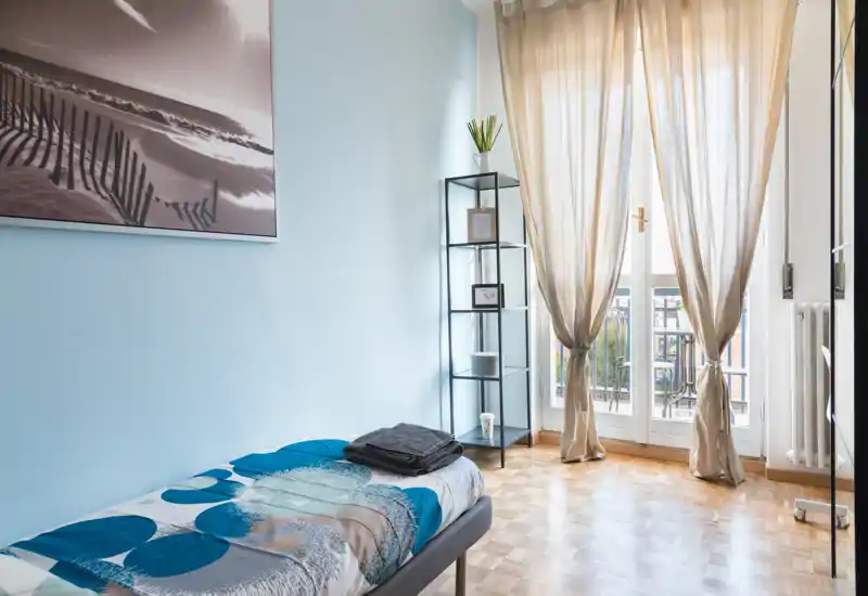 Bright, tidy single bedroom with a neatly made bed, soft blue walls and a balcony door that floods the room with natural light. Minimalist shelving and a framed print create a calm, airy atmosphere ideal for short-term rentals.