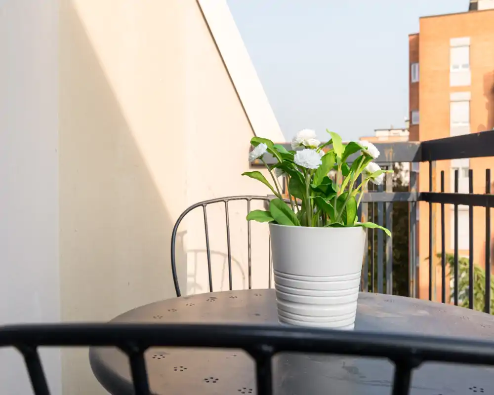 Pequeño balcón soleado con mesa y silla de metal; una maceta blanca con flores aporta un toque fresco y se ven edificios de la ciudad al fondo.
