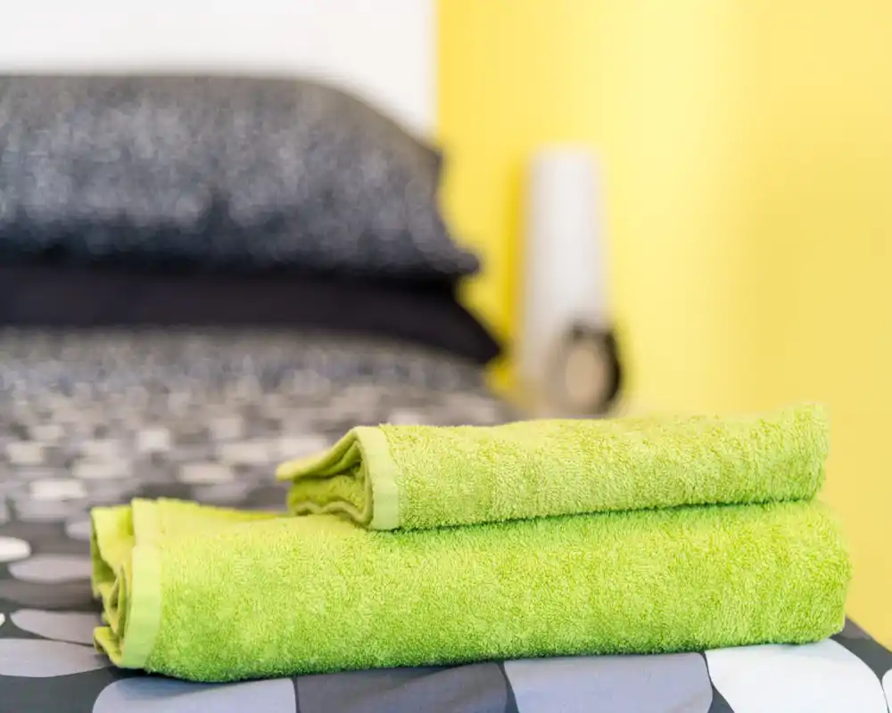 Primer plano de un dormitorio luminoso y bien ambientado: toallas dobladas sobre la cama, ropa de cama estampada y una pared de acento amarilla.