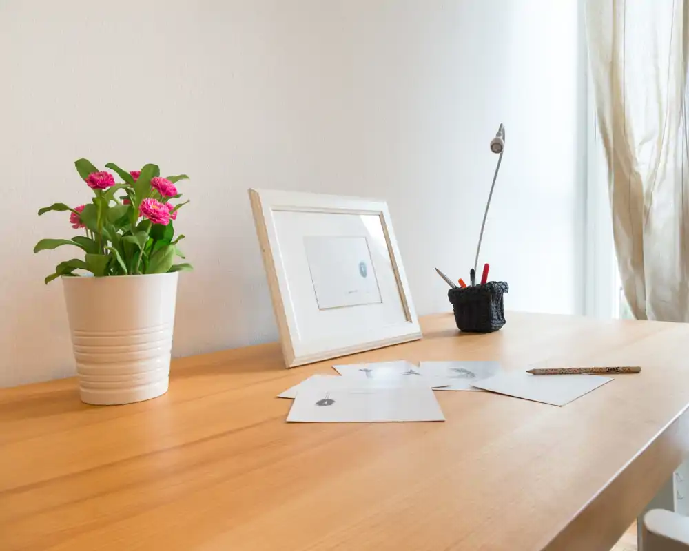 Detalle luminoso y minimalista de un salón con superficie de madera, maceta con flores, marco y pequeña lámpara de escritorio — un rincón tranquilo y bien iluminado para relajarse o trabajar ligeras tareas.