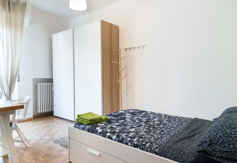 Dormitorio luminoso y ordenado con cama individual, armario y escritorio junto a la ventana; tonos neutros y luz natural que crean un espacio acogedor.