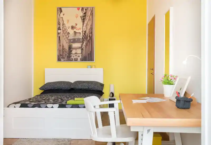 Dormitorio luminoso y ordenado con cama blanca y escritorio de madera junto a la ventana; pared amarilla alegre y decoración moderna que crean un ambiente acogedor.