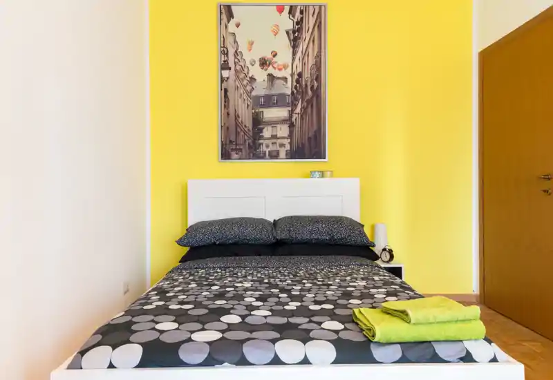 Dormitorio moderno y luminoso con cama doble centrada, ropa de cama estampada y pared de acento amarilla. La habitación está bien iluminada, ordenada y equipada con almohadas, una lámpara de noche y toallas dobladas.